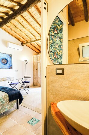 Ein rustikales Interieur mit Holzbalkendecke, einem Essbereich mit Tisch und Stühlen, einem Bett mit floralem Überwurf und einem Badezimmer mit Holzwaschtisch.