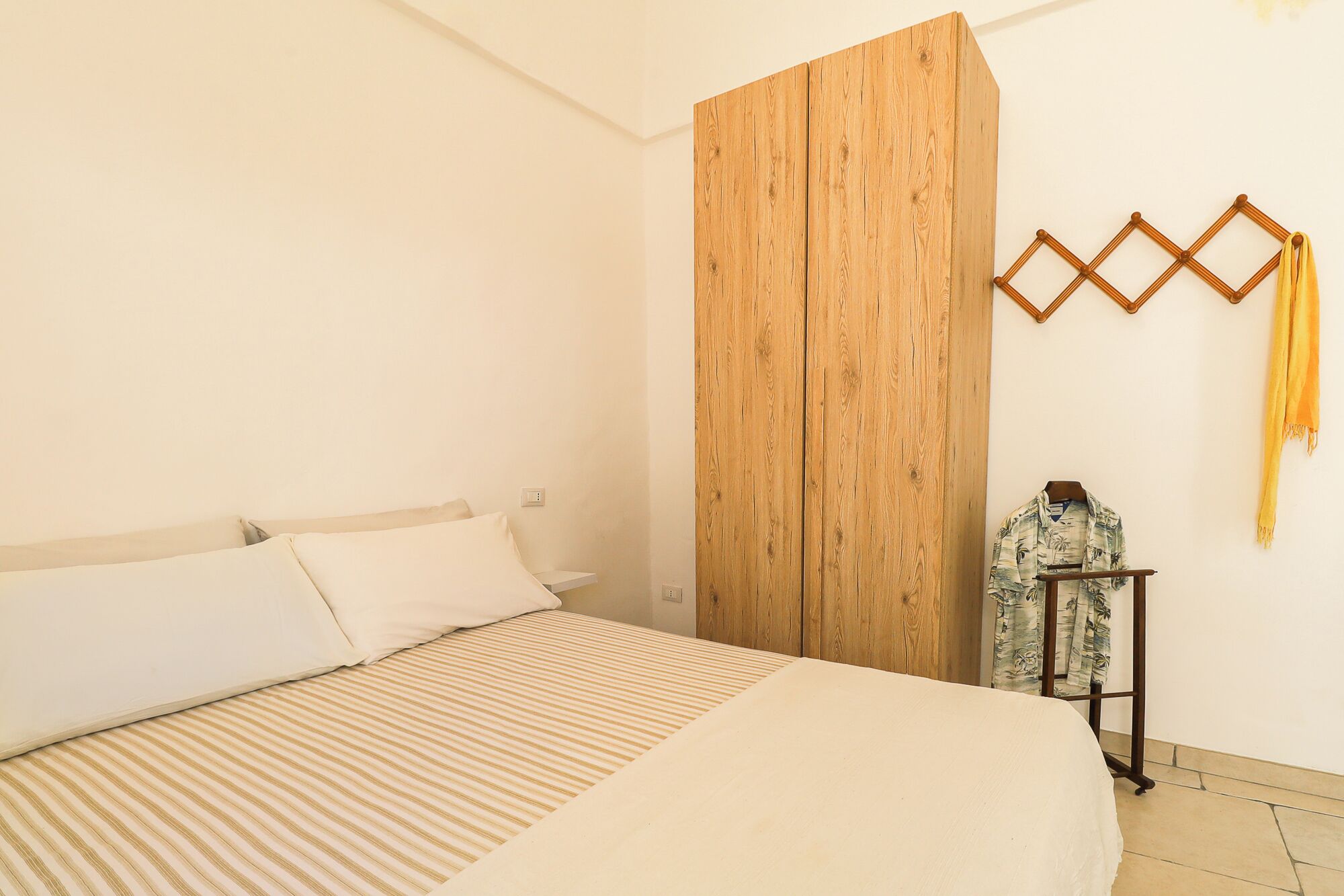 A bedroom with a bed, a wooden wardrobe, a hanging rack with clothes, and a wall-mounted light fixture.
