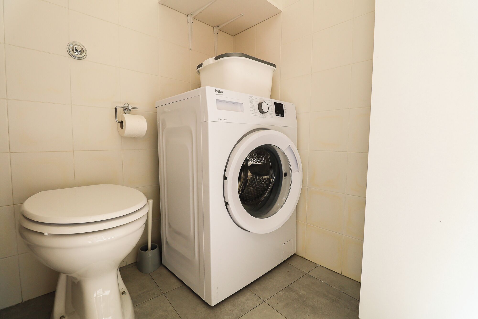 A compact bathroom with a toilet, a washing machine, and a small trash bin.