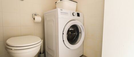 A compact bathroom with a toilet, a washing machine, and a small trash bin.