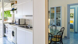 A compact kitchen with a washing machine, a toaster, and a sink. There is a dining area with a table and chairs visible through a glass door. - Casa sul mare Rivabella by Casa di Marco (Rivabella)