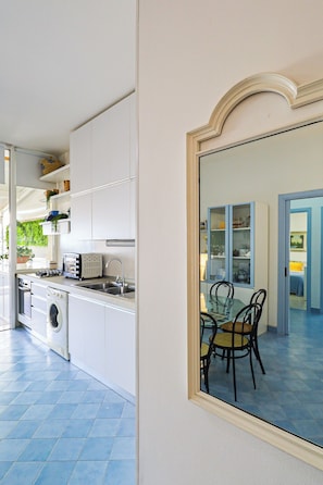 A compact kitchen with a washing machine, a toaster, and a sink. There is a dining area with a table and chairs visible through a glass door. - Casa sul mare Rivabella by Casa di Marco (Rivabella)