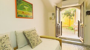 Een kamer met een bed, een kussen met bladmotief, een wandairconditioner en een open deur naar een buitenruimte met een stoel en planten.