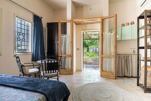 Ein Zimmer mit Bett, Stuhl, Tisch, einem großen Fenster mit Vorhängen, einer Tür zum Balkon und einer Küchenzeile mit Schränken und Spüle.