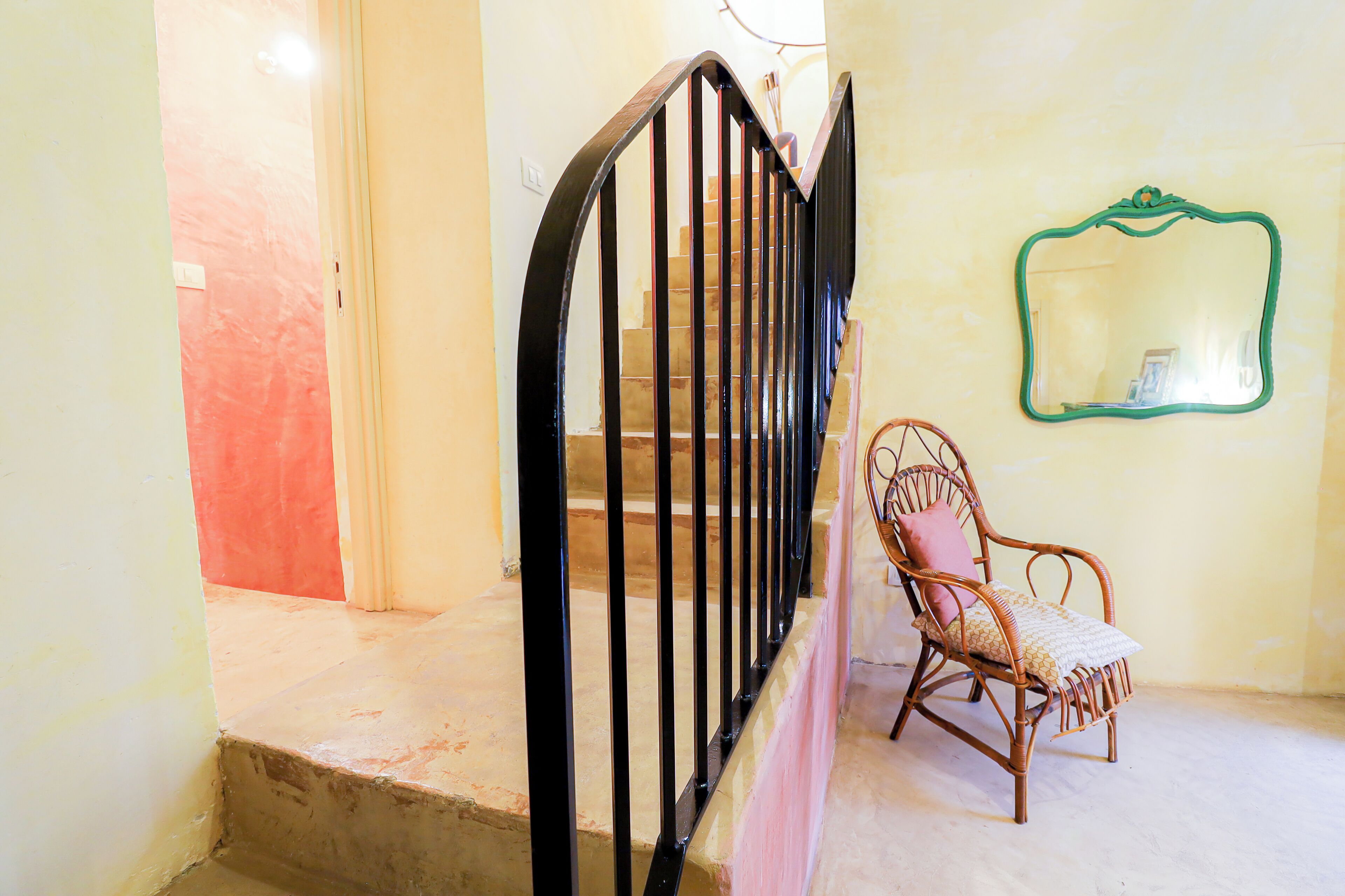 Un escalier avec des rampes noires, une chaise en osier avec un coussin et un miroir au mur.