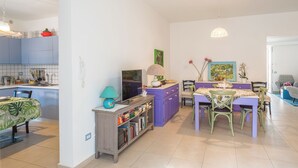 Eine moderne Küche mit blauen Schränken, eine Essecke mit einem violetten Tisch und ein Wohnbereich mit Fernseher und Bücherregalen.