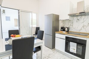 Eine moderne Küche mit Kühlschrank, Backofen und Kaffeemaschine. Ein Esstisch mit Stühlen und eine Schüssel mit Obst sind ebenfalls vorhanden.