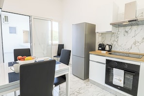 Una cocina moderna con refrigerador, horno y cafetera. Hay una mesa de comedor con sillas y un bol de fruta.