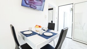 Un'area pranzo con un tavolo bianco apparecchiato per due persone, una ciotola di frutta e un televisore a parete che mostra un'immagine di un telefono rosa.