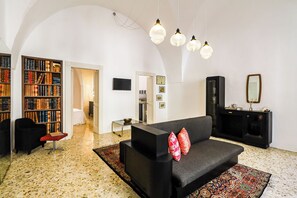 Een ruime woonkamer met een zwarte bank, een salontafel en een boekenplank vol boeken. Er is een televisie aan de muur bevestigd en een spiegel boven een zwart kastje.
