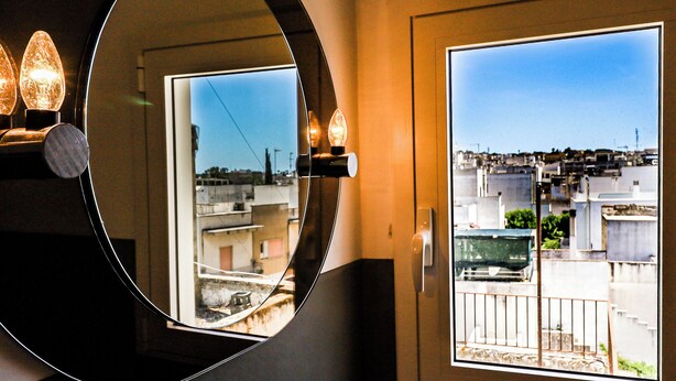 A room with a large mirror reflecting a view of a cityscape through a window. - Palazzo Frascà by Casa di Marco (Parabita)