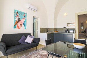 Un salon moderne avec un canapé gris foncé, une table basse en verre et un tapis à motifs. On aperçoit au fond une cuisine avec des meubles gris et un petit îlot central.