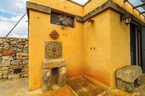 Un coin d’un bâtiment avec un mur en pierre, une petite fenêtre ornée d’une plante, une gouttière et un banc en pierre.
