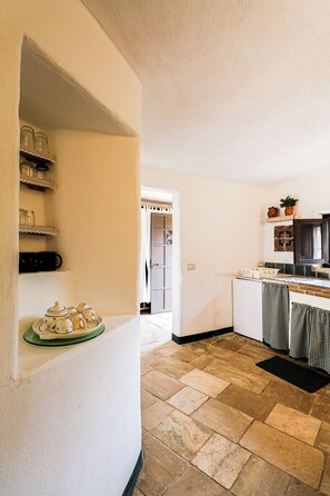 Una cocina con piso de piedra, paredes blancas y un acceso a otra habitación.