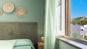 Ein Schlafzimmer mit Bett, Nachttisch mit Lampe, Fenster mit Blick auf Gebäude und Grünflächen sowie Wanddekorationen aus gewebten Textilien.