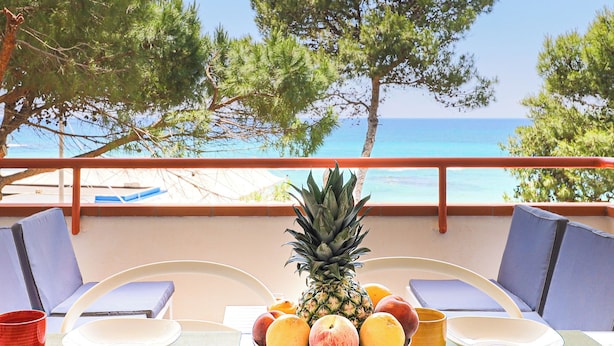 Ein Tisch mit Obstschale, Tellern und Tassen mit Blick auf einen Strand, Bäume und das Meer.
