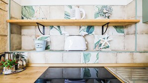 Eine Küche mit einem Holzbodenregal, auf dem ein Wasserkocher, ein Toaster und ein dekorativer Seestern stehen. Darunter befindet sich ein Spülbecken mit einem blauen Eimer und einem Blumentopf.