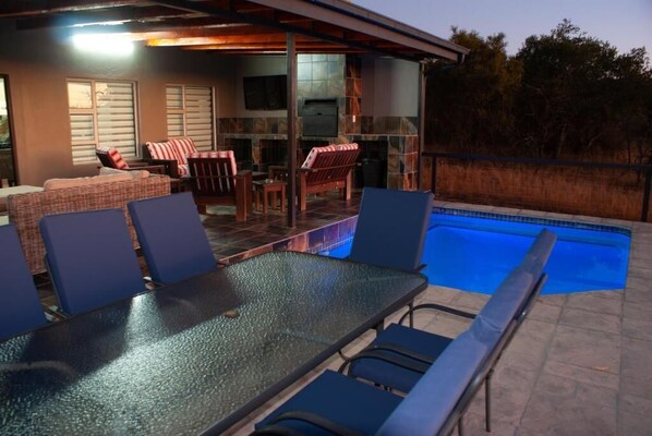 An outdoor pool area with lounge chairs and a table, surrounded by a covered patio with seating. - Casa de la Presa 1 (Polokwane)