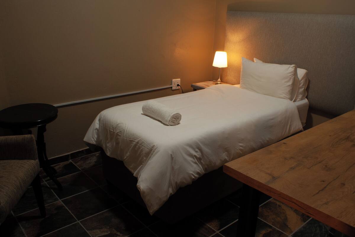 Une chambre d’hôtel avec un lit, une table de chevet avec une lampe, une table en bois et une chaise.