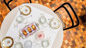 Ein runder Tisch, gedeckt für eine Mahlzeit, mit tellerförmigen Dekor in floralem Muster, einer Servierplatte mit Brot in der Mitte und einer pinkfarbenen Teekanne.