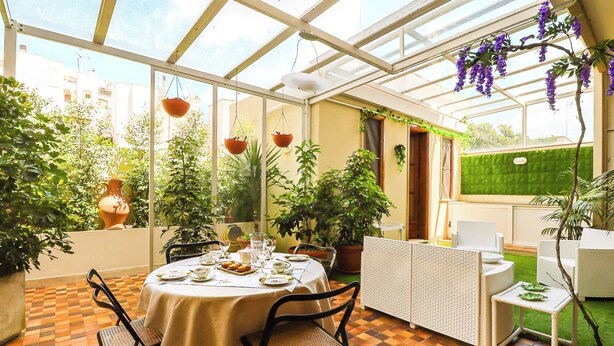 Ein Wintergarten mit Esstisch, gedeckt für vier Personen, umgeben von Blumentöpfen und Blick auf die Stadtlandschaft.