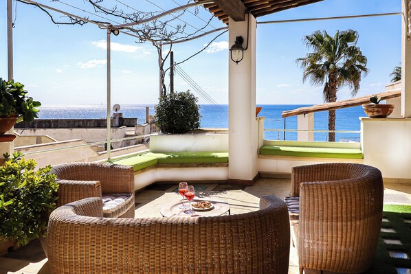 An outdoor patio with wicker furniture, a table with a glass of red wine, and a view of the sea.