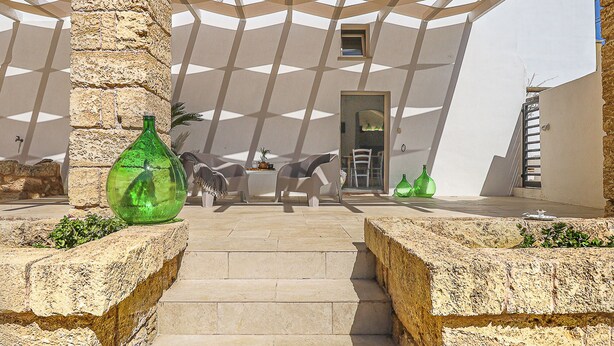 Un espacio exterior moderno con una estructura de techo geométrica, escalones de piedra y una puerta que da a un espacio interior.