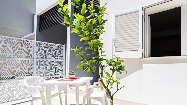 Un área de estar al aire libre con una pequeña mesa y sillas, una valla blanca decorativa y una ventana con persianas cerradas.