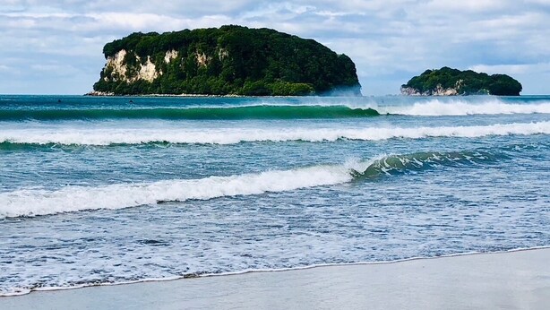 Uma praia com ondas quebrando na costa e duas ilhas verdes ao longe.