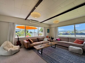 A modern living room with a sofa, a coffee table, and a guitar. There are large windows with an umbrella outside. - Last Minute Beach Hop (Whangamatā)