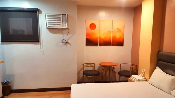 Une chambre d’hôtel comprenant un lit, une petite table avec deux chaises, un appareil de climatisation et une œuvre d’art fixée au mur représentant un coucher de soleil sur des montagnes.