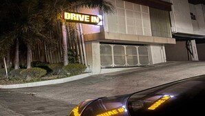 Un bâtiment moderne avec une enseigne « Drive In », une porte de garage et une allée bordée de palmiers.