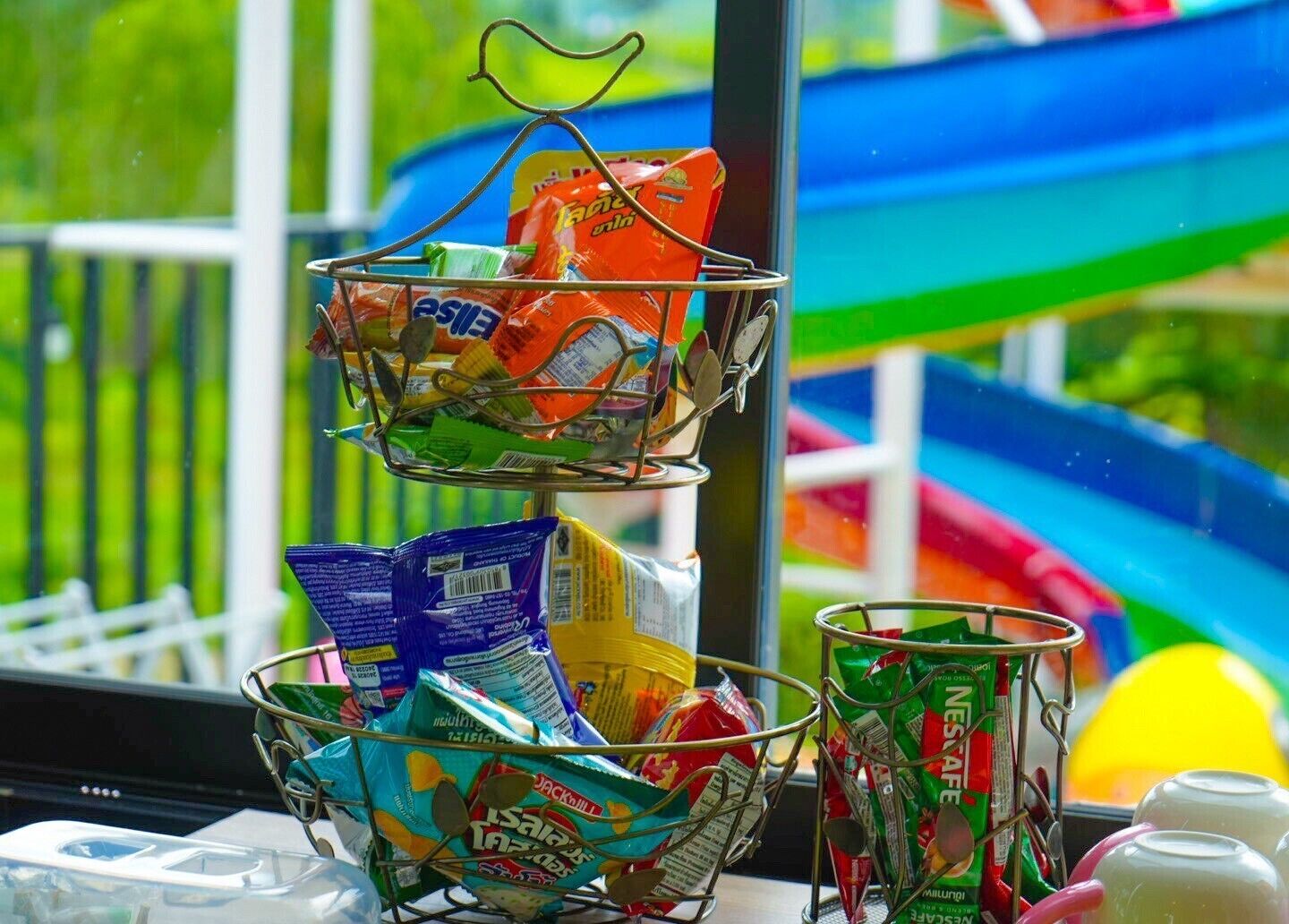 A window with a view of a colorful slide, a metal rack with snack bags, and a glass with a beverage.