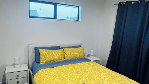 A bedroom with a bed, two bedside tables, a window, and a curtain. - Room-room With En-suite and Kitchenette (Kaiapoi)