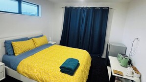 A bedroom with a bed, a nightstand, a desk with a kettle, and a window with curtains. - Room-room With En-suite and Kitchenette (Kaiapoi)
