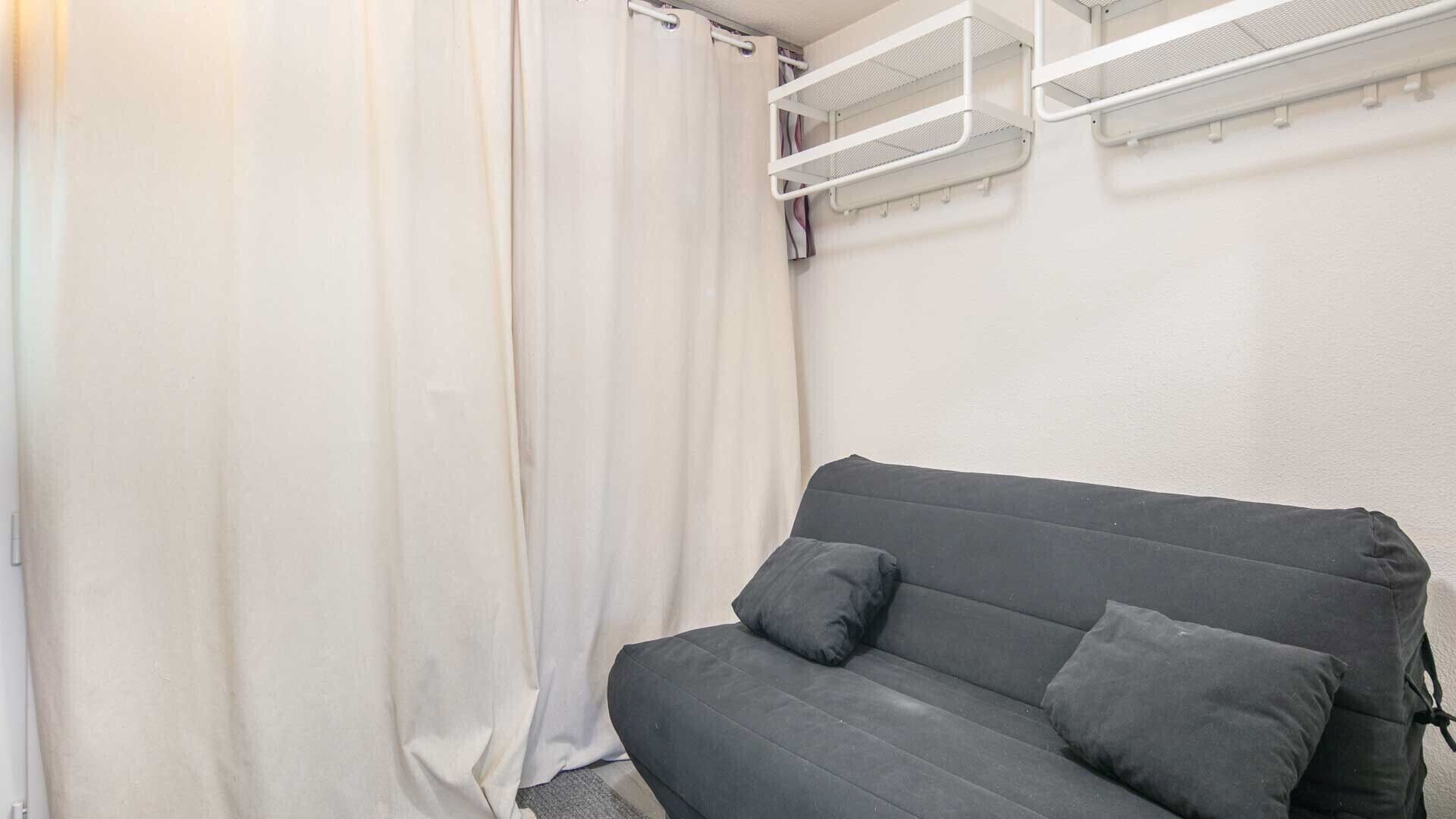 En grå soffa med två kuddar i ett rum med vita väggar och en vit gardin.