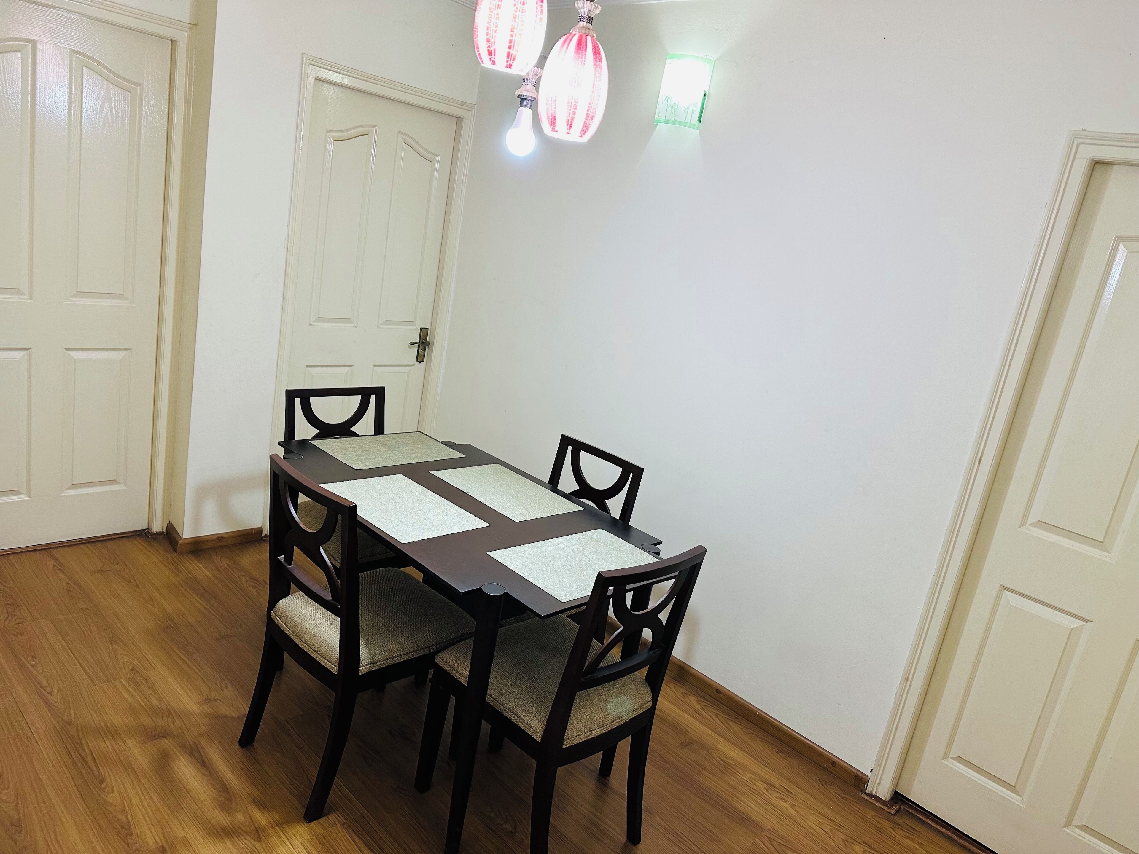 Un comedor con mesa de madera y cuatro sillas, paredes blancas y dos puertas.