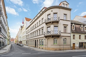 A street view of a building with a curved facade, multiple windows, and a balcony with a decorative railing.