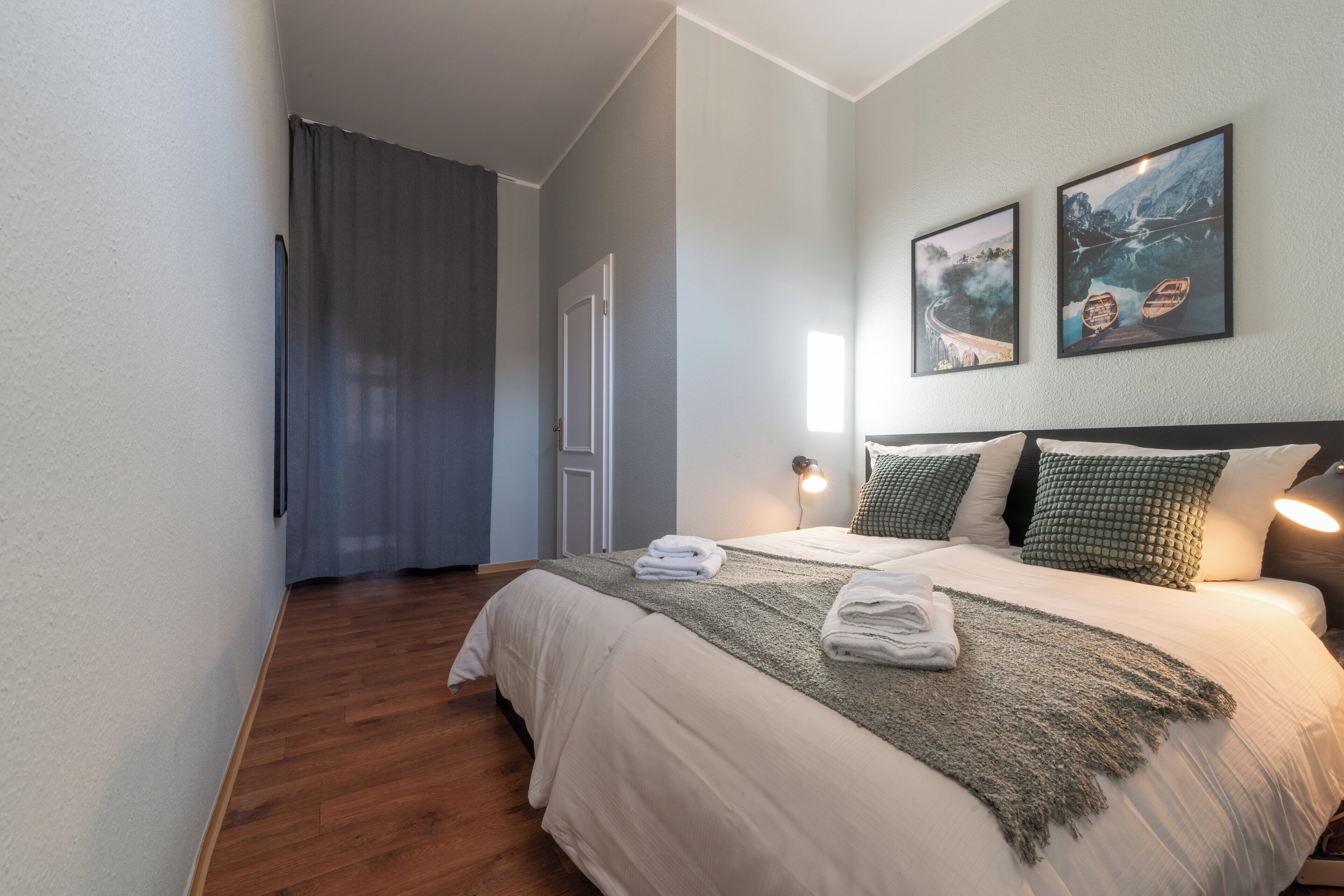 Een netjes opgemaakt bed met een witte dekbedovertrek en een groene plaid, een houten vloer, een grijze gordijn en twee ingelijste foto’s aan de muur.