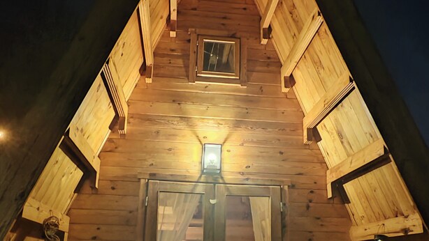 A wooden cabin with a pitched roof, a small window, and a door with curtains. Inside, there is a table and two chairs.