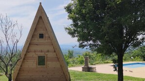 A wooden triangular cabin with two windows, surrounded by a grassy area and a swimming pool.