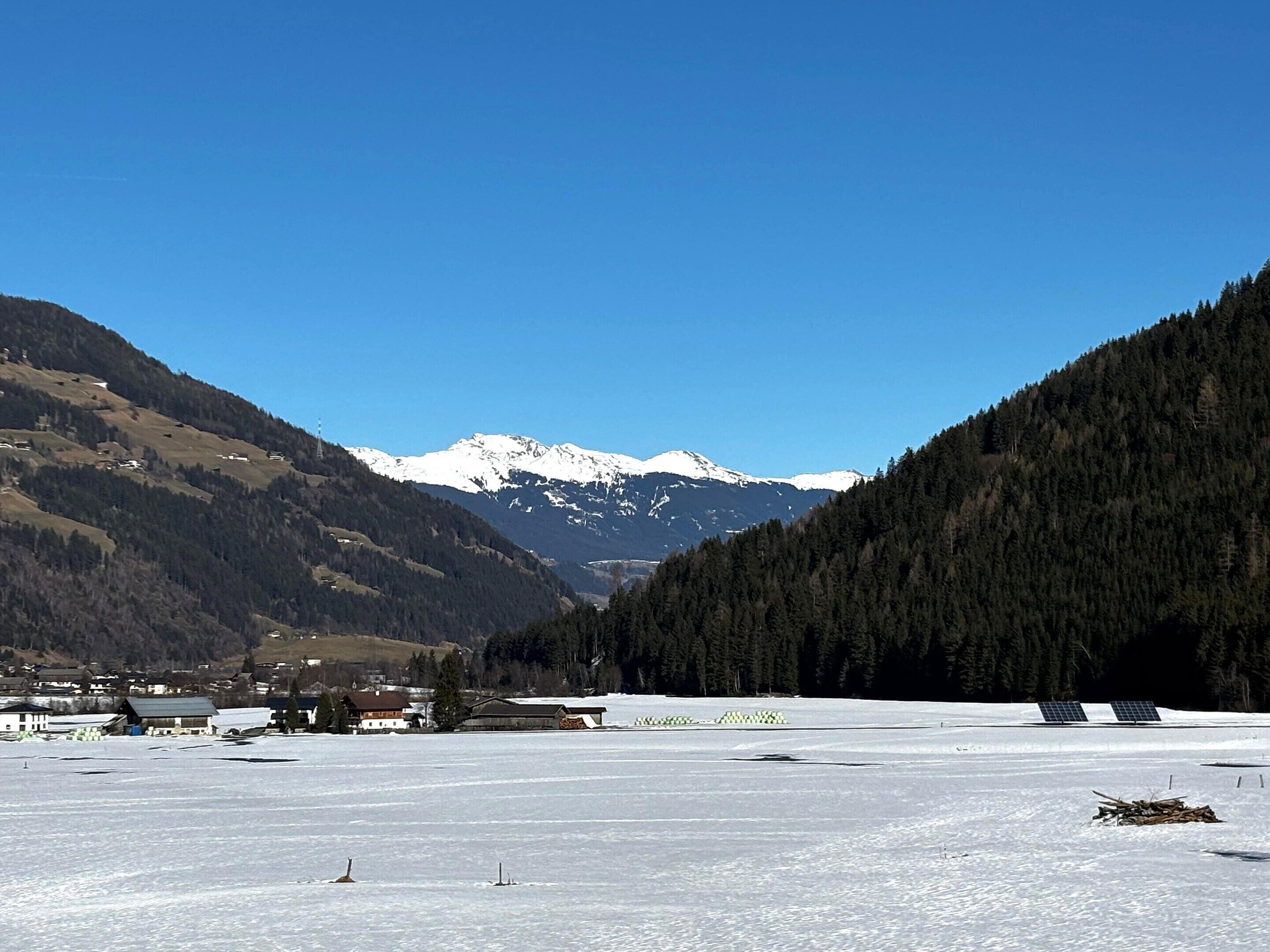 A snowy landscape with a mountain range in the background, a clear sky, and a few buildings in the foreground.