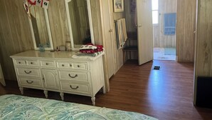 A bedroom with a wooden floor, a white dresser with multiple drawers, a bed with a patterned bedspread, and a door leading to another room.