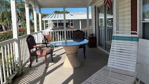 A covered porch with a table and chairs, an American flag, and a view of a house with a white fence and red flowers.
