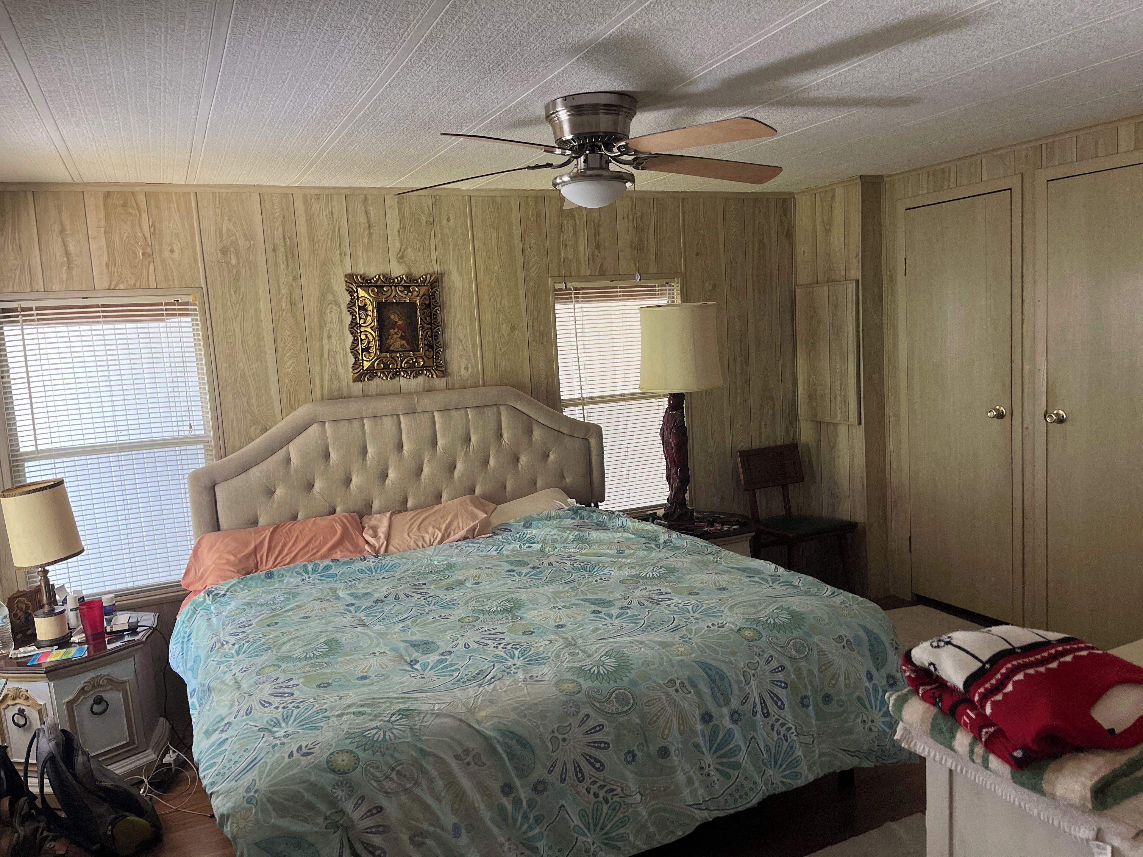A bedroom with a tufted headboard, a bed with patterned bedding, a ceiling fan, a nightstand with a lamp, and a wardrobe.