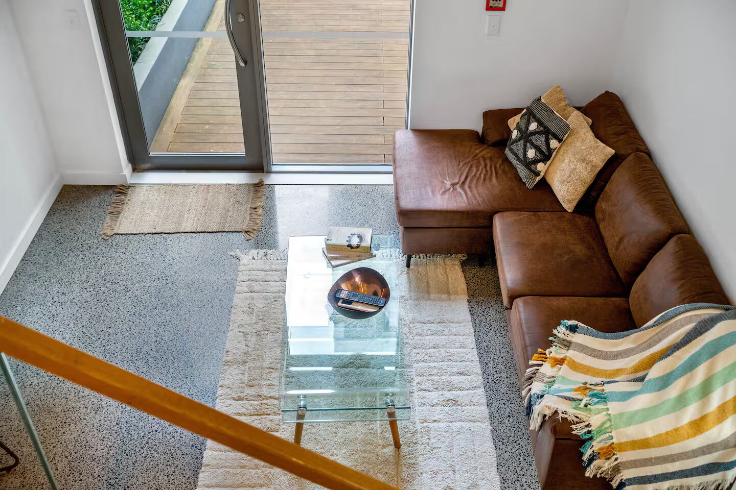 A modern living room with a glass coffee table, a brown leather sofa, and a view through a glass door to a wooden deck.