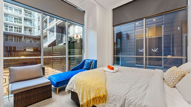 A modern hotel room with a large bed, a blue armchair, a small sofa, and a view of a building with balconies. - 2BR Loft |A/C, Netflix, Private Deck| Walk to City (Auckland Central)