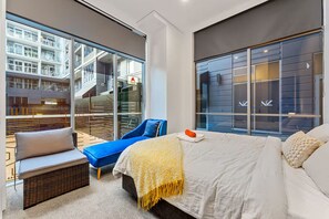 A modern hotel room with a large bed, a blue armchair, a small sofa, and a view of a building with balconies. - 2BR Loft |A/C, Netflix, Private Deck| Walk to City (Auckland Central)