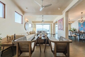 A spacious living area with a high ceiling, a dining table with chairs, and a view to the outside.