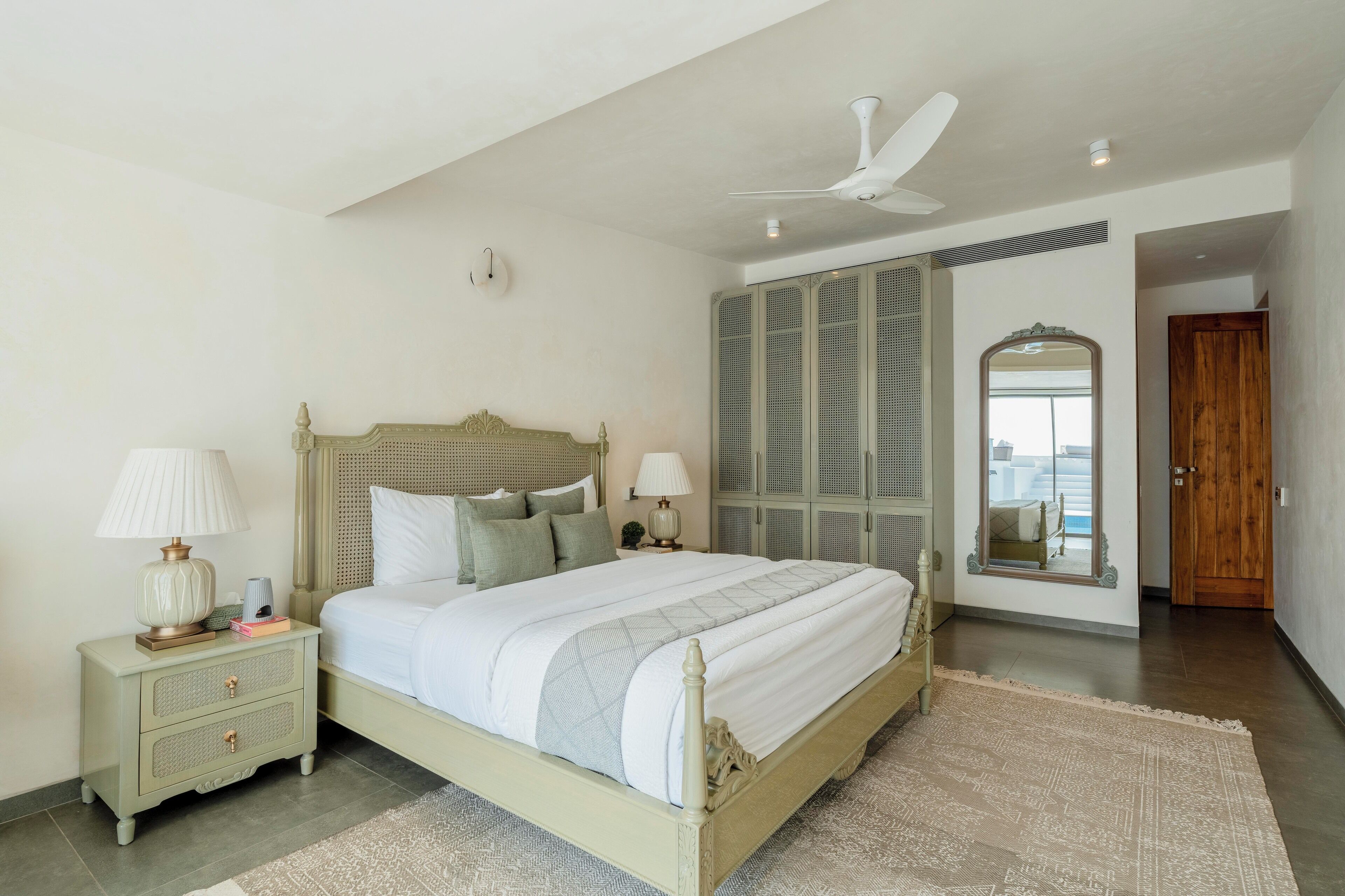 A bedroom with a bed, nightstands, a ceiling fan, a large mirror, and a wooden door.